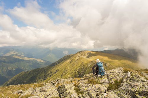 Free Back view anonymous male traveler with backpack sitting on grassy hilltop and admiring majestic landscape of mountainous terrain Stock Photo