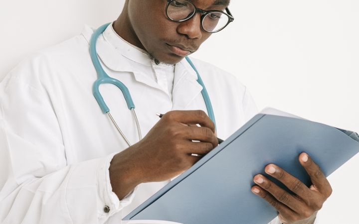 A doctor wearing eyeglasses writes on a medical chart indoors, displaying professional focus.