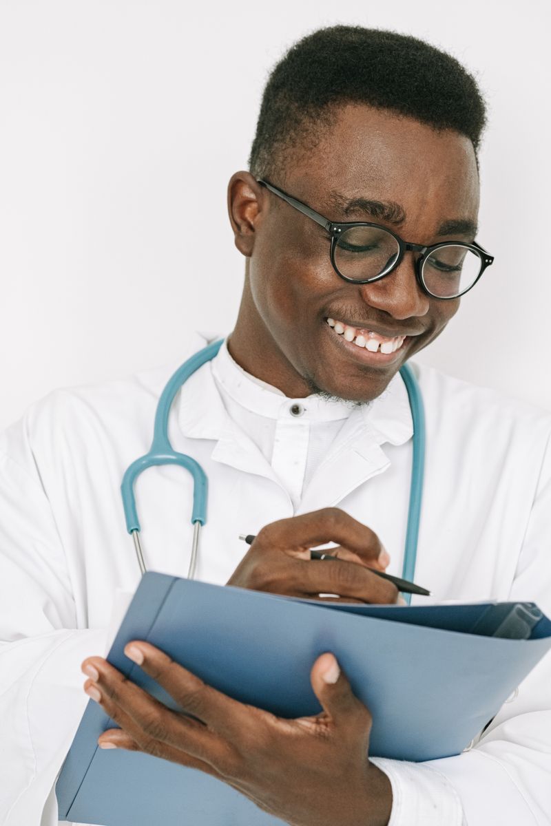 A Black physician in a white coat and stethoscope writes on a chart, the kind of nephrology visit where heavy protein in the urine and early kidney damage get caught.