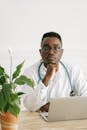 一位非裔美国医生坐在办公桌前，室内环境宁静，他戴着听诊器，手里拿着笔记本电脑和一盆植物