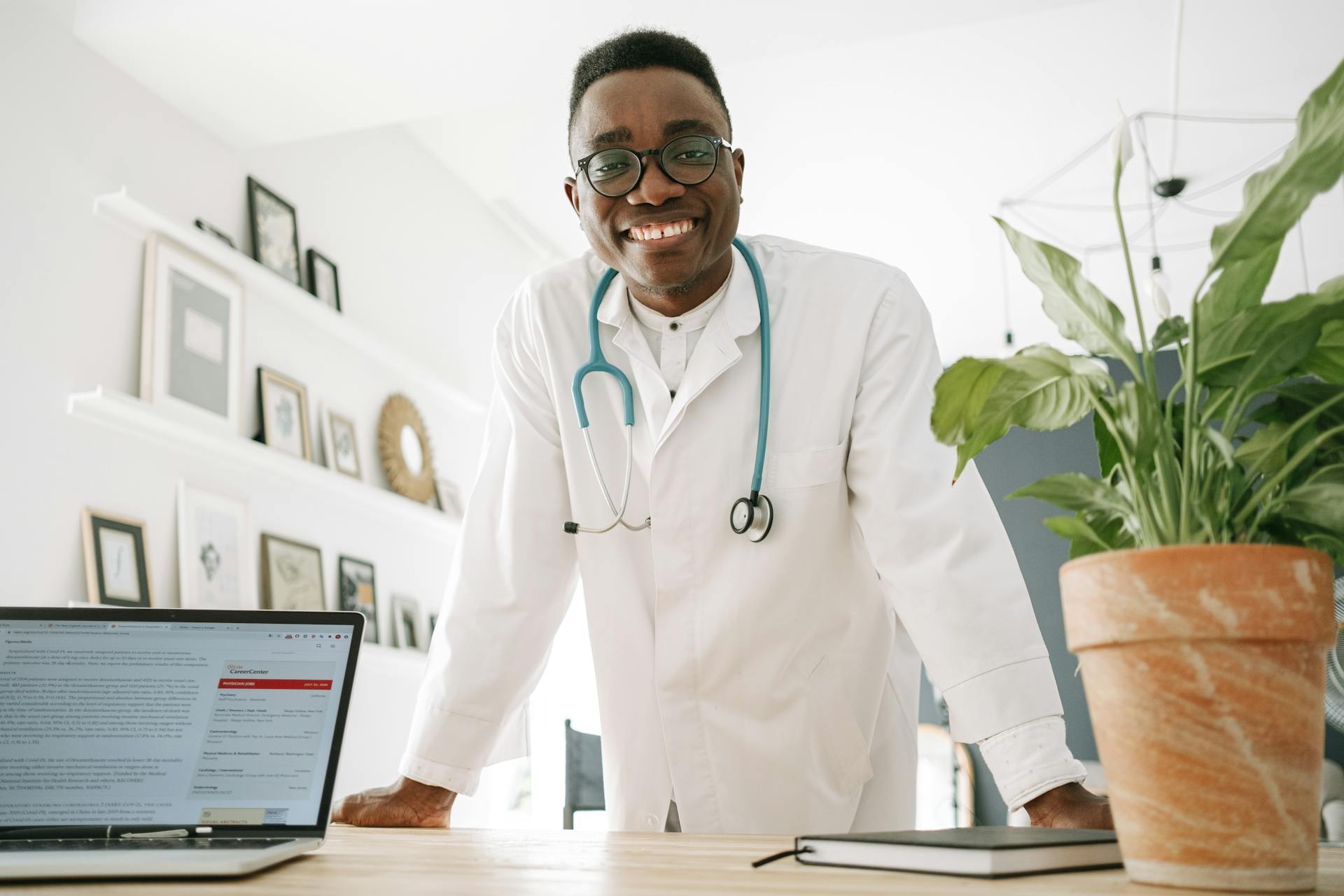 Smiling doctor in white coat and stethoscope leaning on desk in a modern workspace.