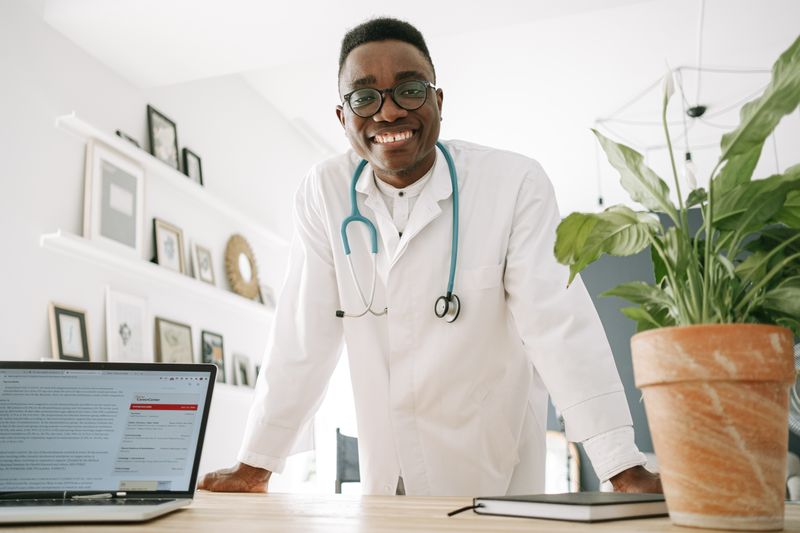 A Black male doctor in a white coat with a stethoscope smiles warmly while leaning on a desk in a bright, modern office.