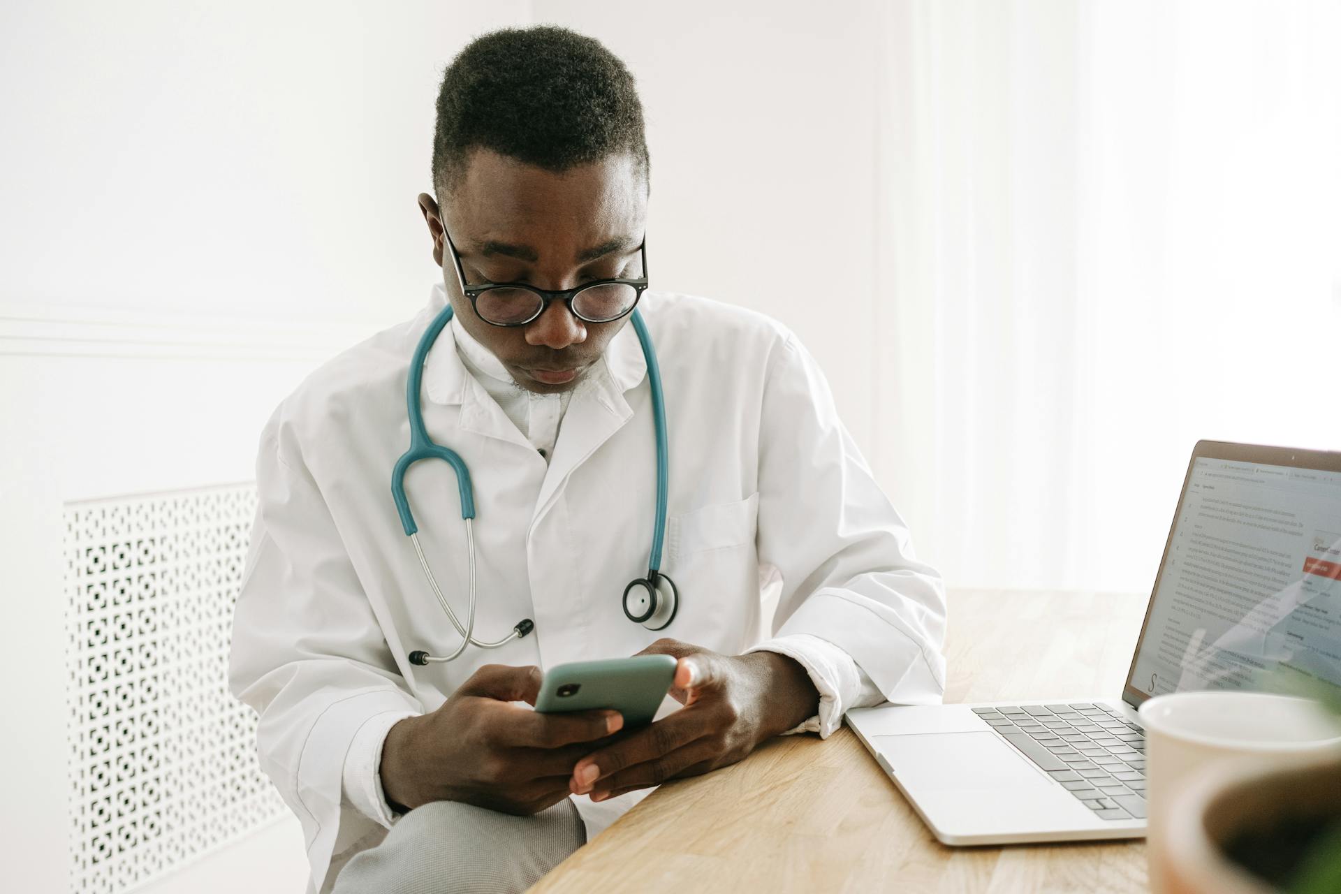 Doctor in a medical office checking a smartphone with a laptop on the table, wearing eyeglasses and stethoscope.