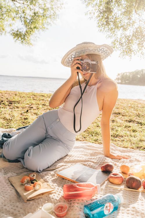 Free A woman in a hat enjoys photography and a picnic by the serene lakeside on a sunny day. Stock Photo
