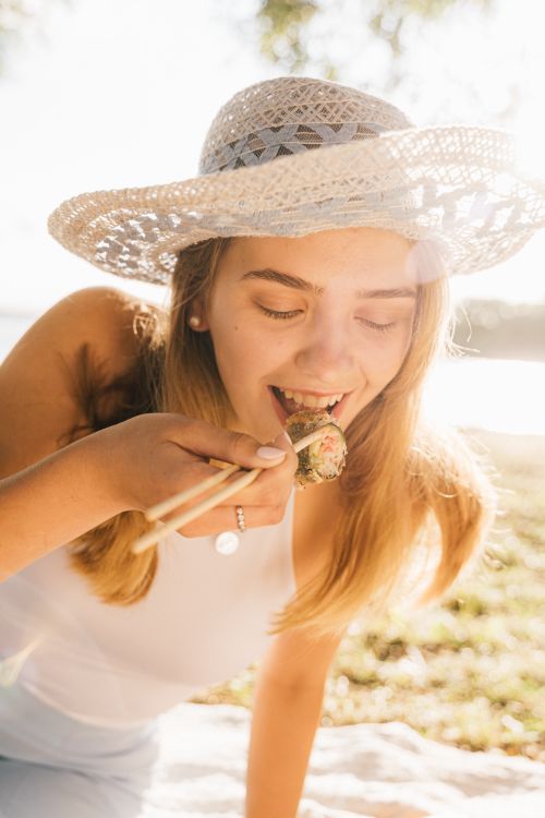 Free A woman in a hat enjoys sushi with chopsticks during a sunny park picnic. Stock Photo