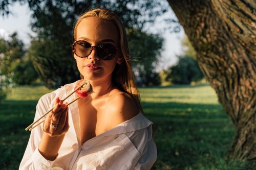 Free Young woman in sunglasses enjoys sushi with chopsticks during a summer picnic in the park. Stock Photo