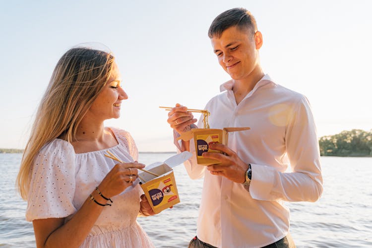 A Man And A Woman Eating Noodles From The Take Out Carton Box