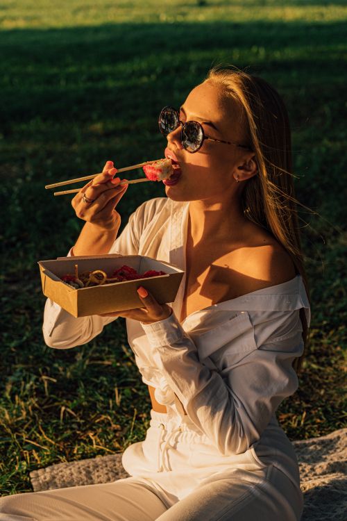 Free Woman in white clothes enjoying sushi with chopsticks during a sunny picnic in the park. Stock Photo