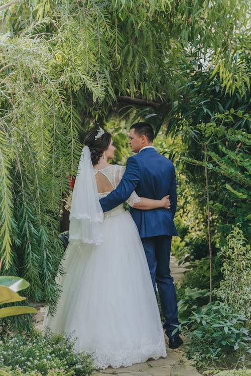 Full body of young couple in wedding clothes hugging and looking at each other in green garden