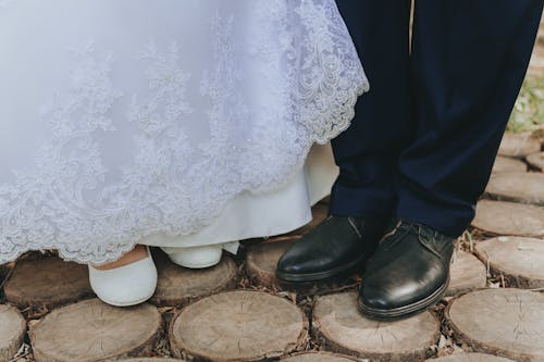 From above of crop bride in white wedding shoes and groom in elegant boots standing on wooden walkway