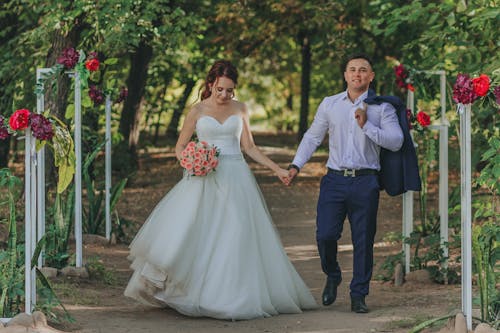 Young stylish groom and bride in white dress with flower bouquet holding hands while strolling on walkway between trees