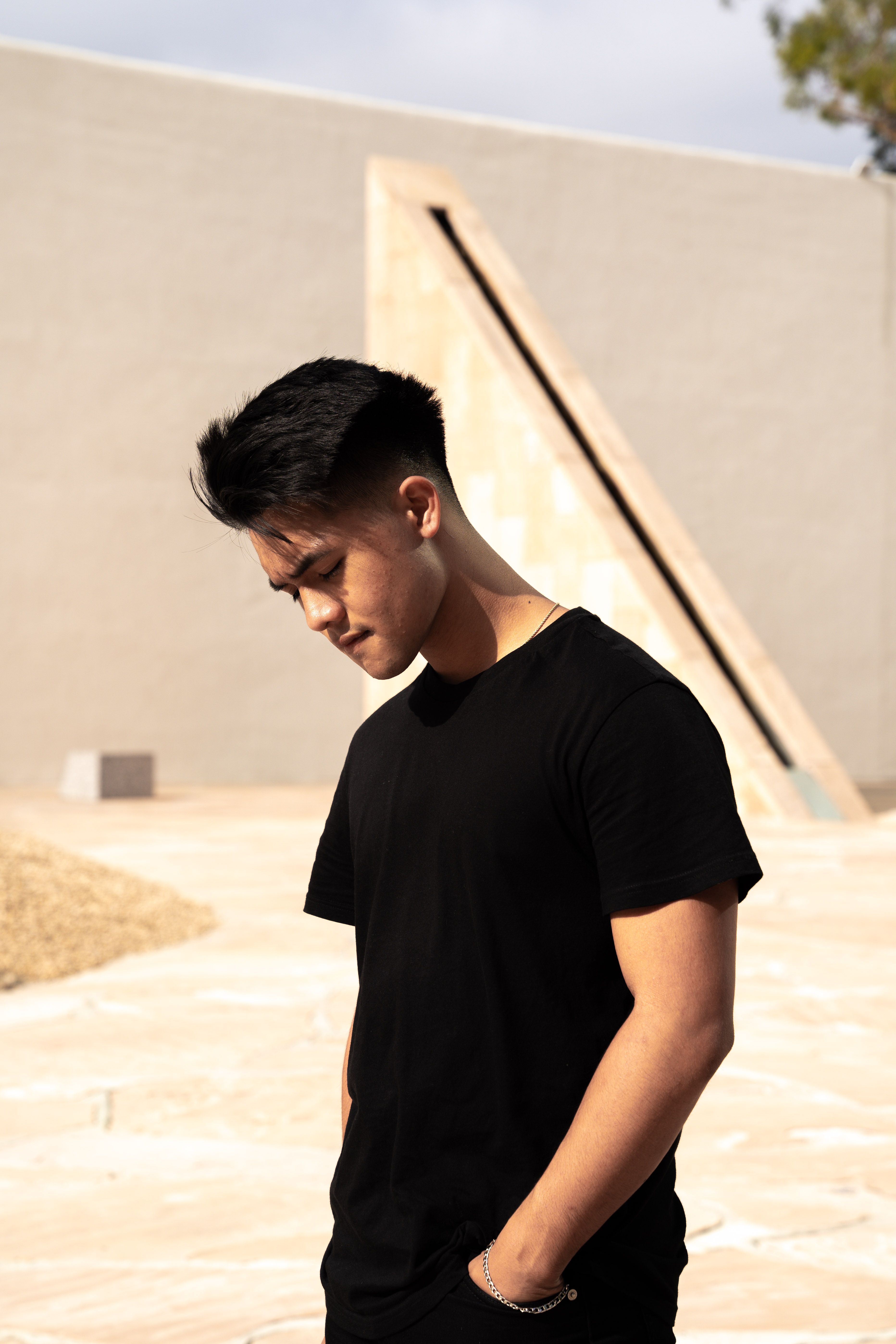Young man in black shirt standing outdoors with hand in pocket, thoughtful pose.