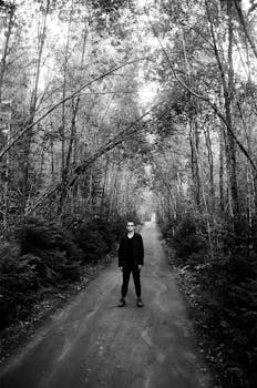 Black and white photo of a man standing alone on a forest pathway, surrounded by tall trees.