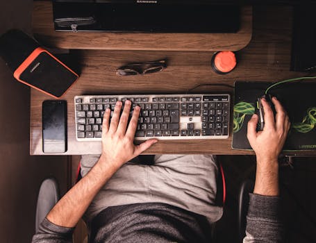 A top-down view of a man using a desktop computer with various electronics on a wooden desk.