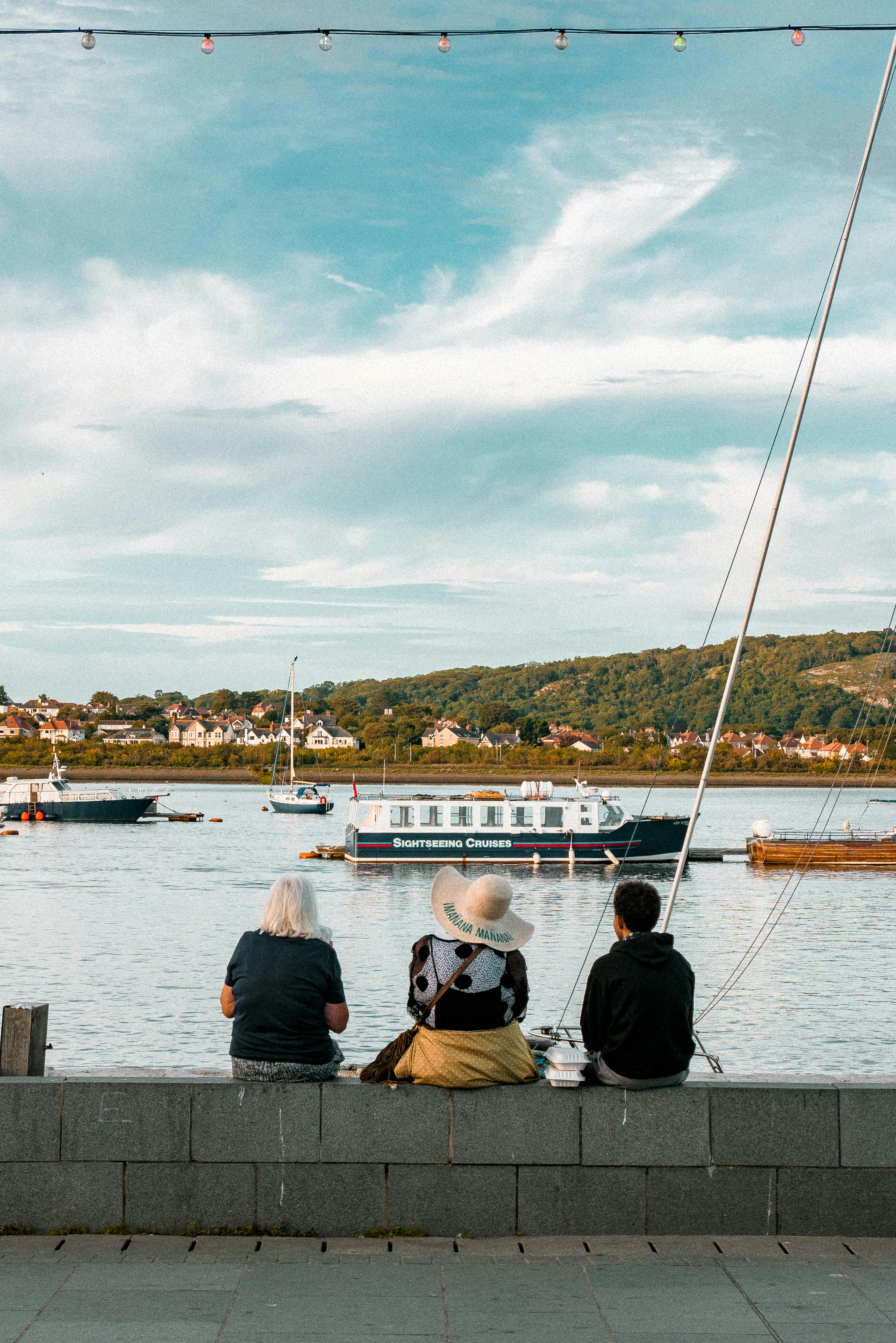 Back view of people in casual clothes sitting on small fence near water and enjoying picturesque view of floating boats
