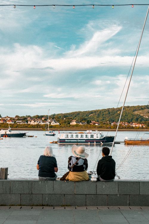 Free Back view of people in casual clothes sitting on small fence near water and enjoying picturesque view of floating boats Stock Photo