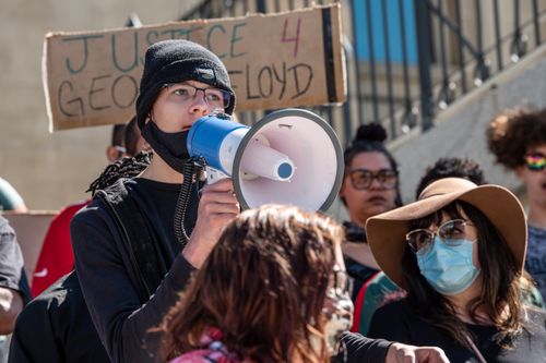 Free Social justice warrior speaking into loudspeaker near anonymous people in masks during strike on street in back lit Stock Photo