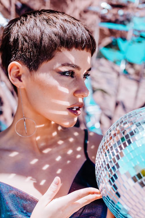 Charming female with bright glitter from silver disco ball looking into distance while standing on foil background