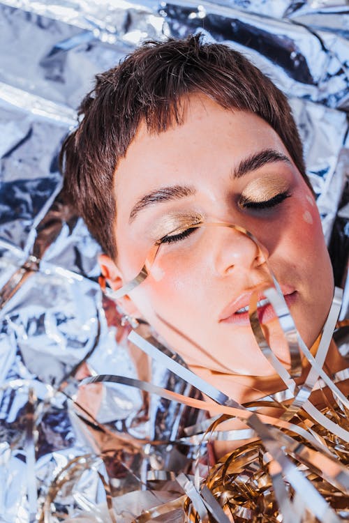 From above headshot of tranquil female with makeup and closed eyes lying on foil covered with decorative silver tinsel