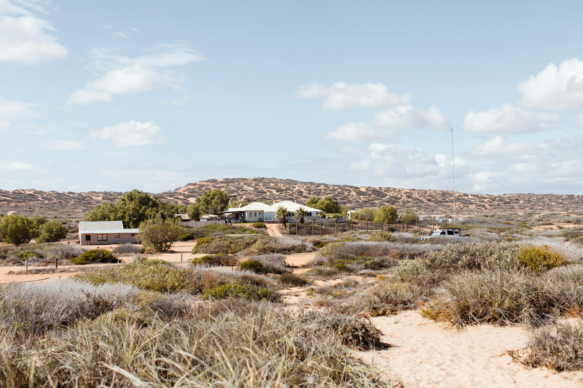 Distant buildings located on dry area with green shrubs against hills in semidesert terrain in rural settlement against cloudy sky