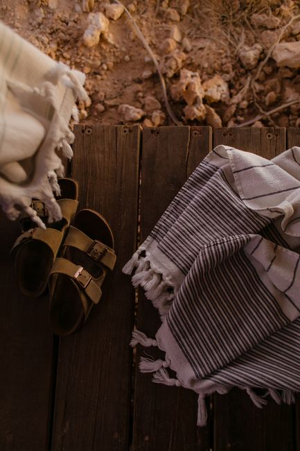 Top view of crumpled fabrics with fringe and leather footwear on wooden planks above sandy land