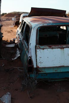 一辆生锈的白蓝相间老式汽车，其损坏的细节被置于沙地上，阴影中可见干燥的植物和金属垃圾