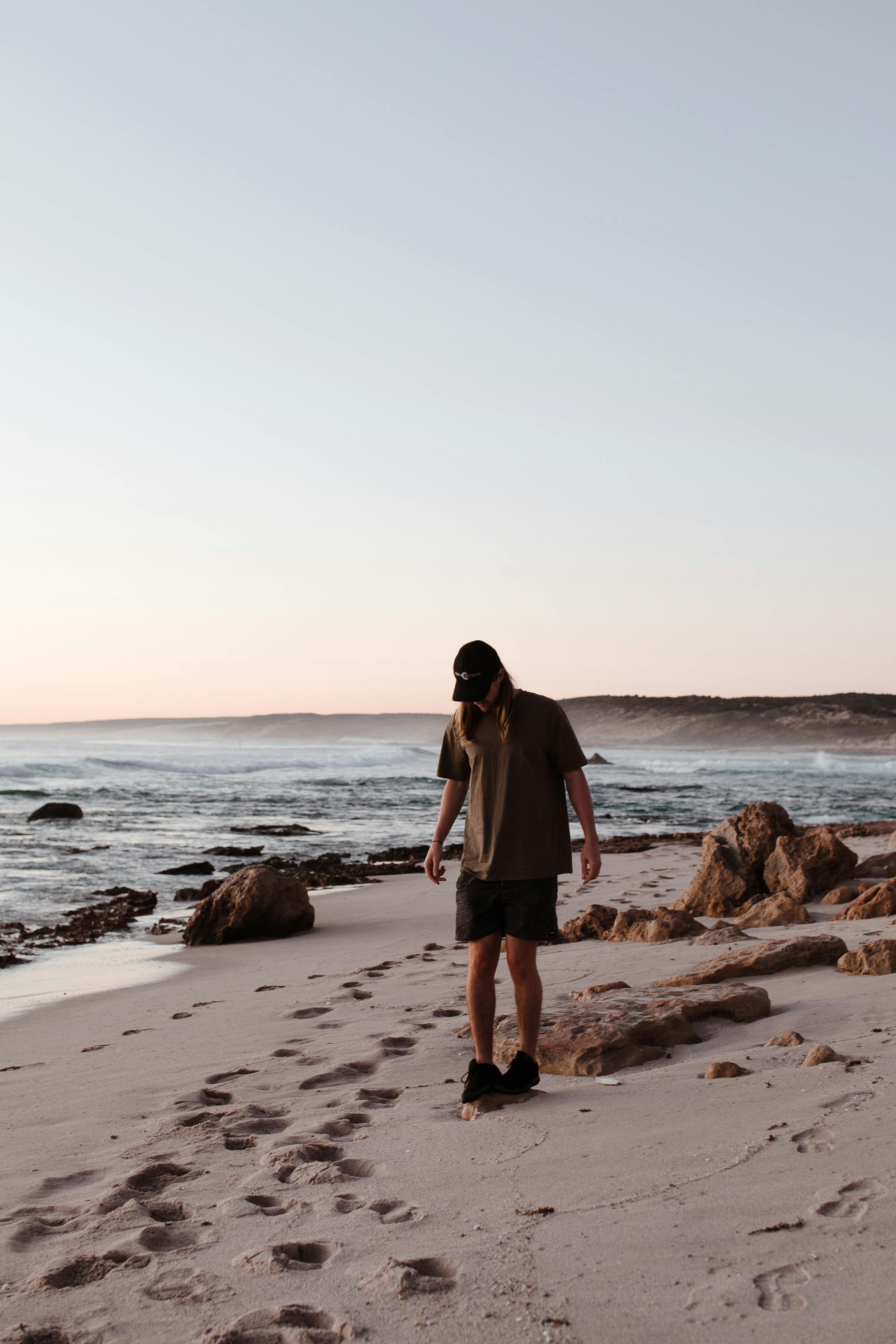 Anonymous male with long hair in casual clothes and cap standing on sandy beach near wavy ocean under cloudless blue sky