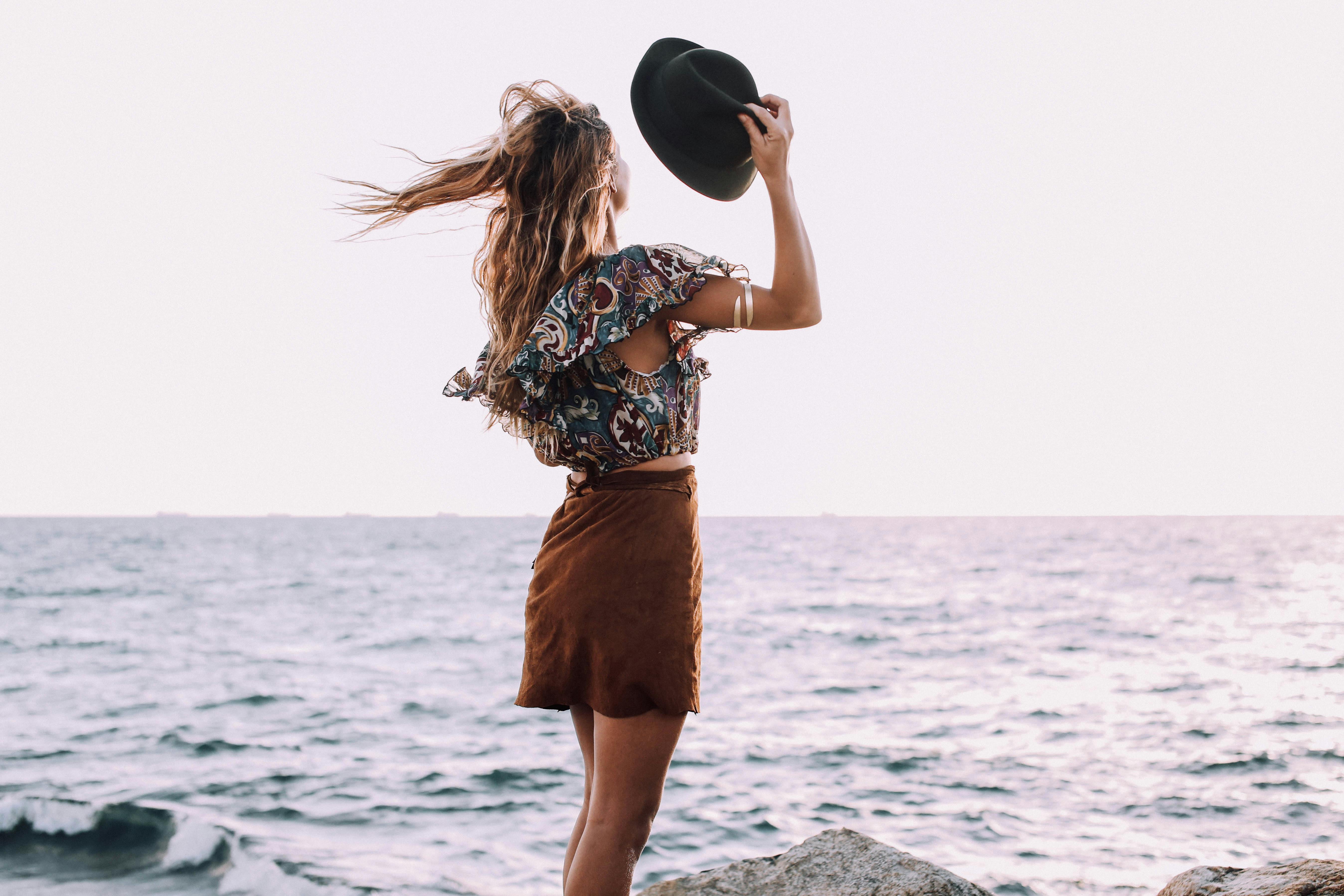 Back view of anonymous woman in stylish outfit standing with black hat in hand on shore near waving sea under cloudless sky in daytime