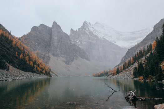 宁静的湖泊四周环绕着秋天的树木，以及晴朗天空下白雪皑皑的山脉