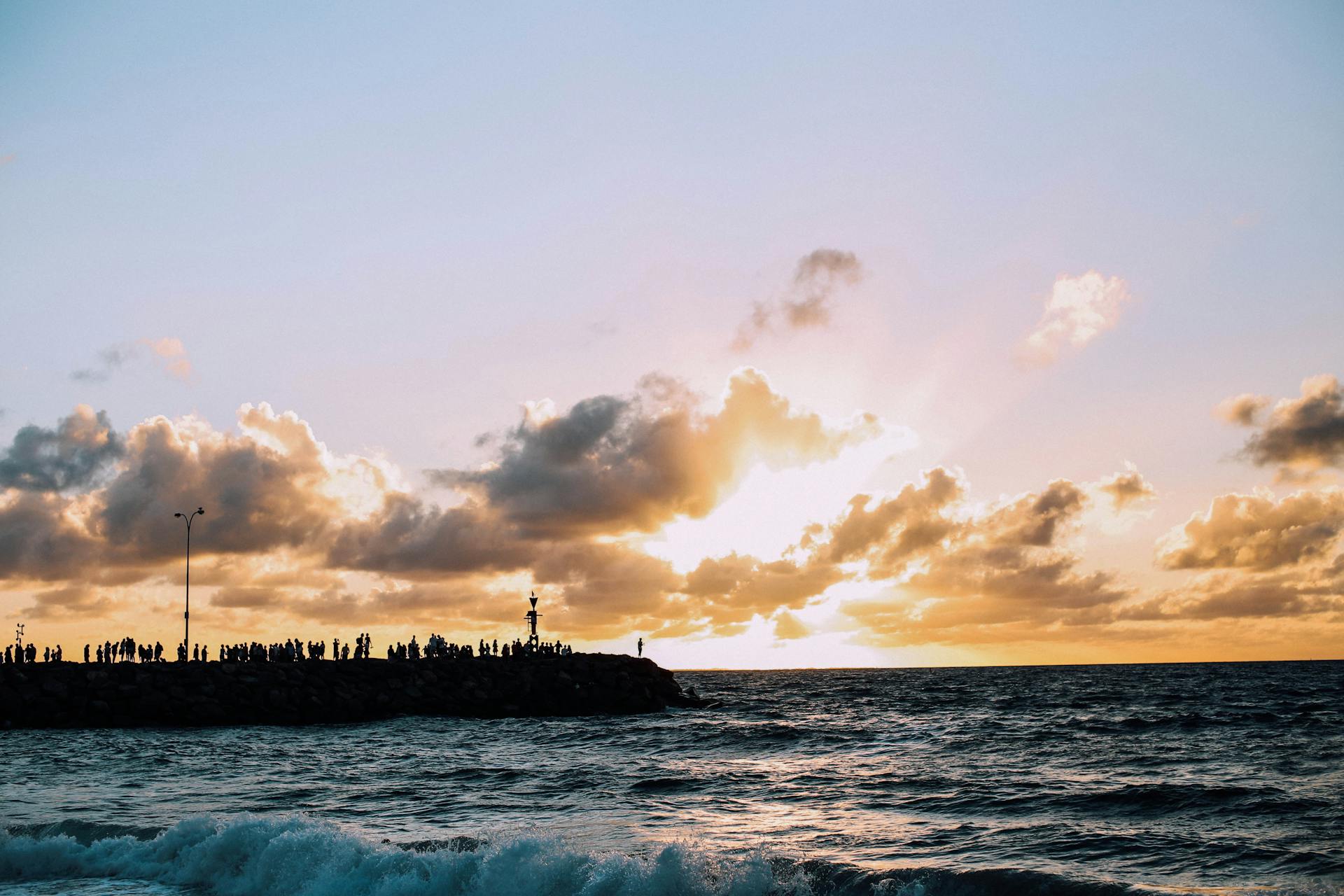 Silhouettes of people standing on embankment of sea with splashing waves under clouds at sunset