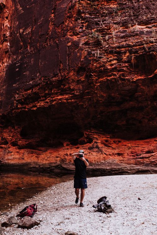 Free Full length of faceless man traveler in casual clothes walking near rocky rough mountains and water while taking picture on photo camera in daylight Stock Photo