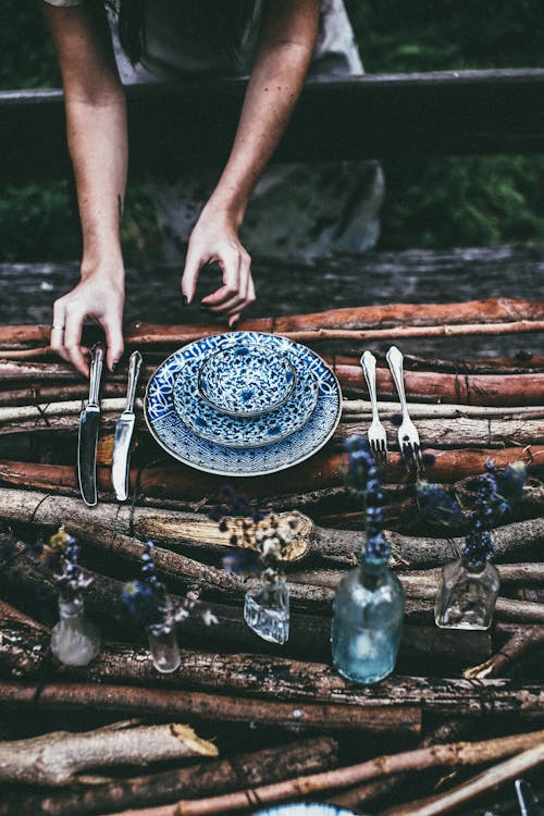 High angle of crop anonymous female putting utensil on wooden surface with plates and forks near knifes and glass bottles in daytime in nature