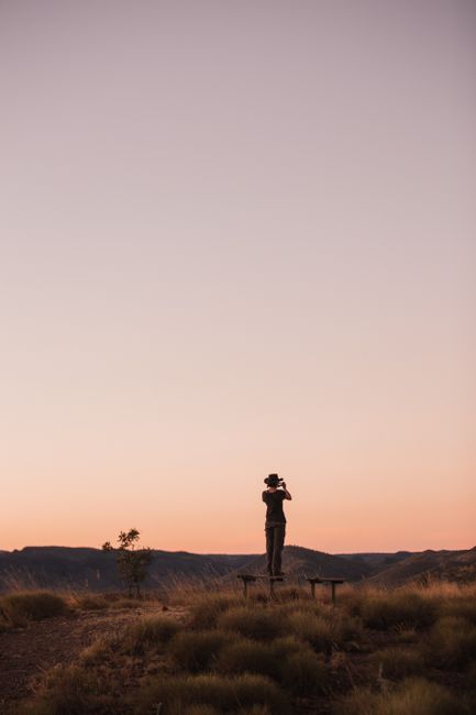 Back view full length of anonymous woman in hat and casual outfit standing on bench in field near green grass and taking photo on mobile of mountains under cloudless sky