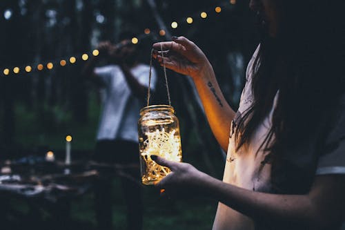 Crop anonymous female with long dark hair in casual clothes standing with glass jar with glowing garland in nature in evening time