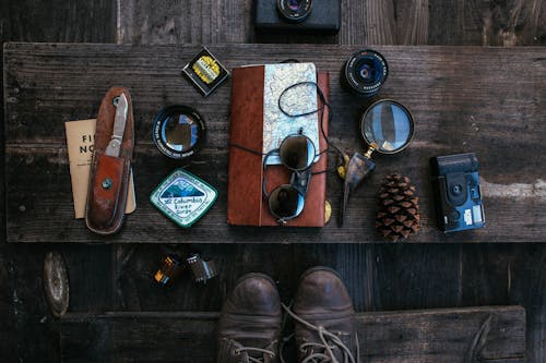 Overhead of vintage photo camera near leather notebook with map and knife near camera roll and lenses near sunglasses placed on table near boots