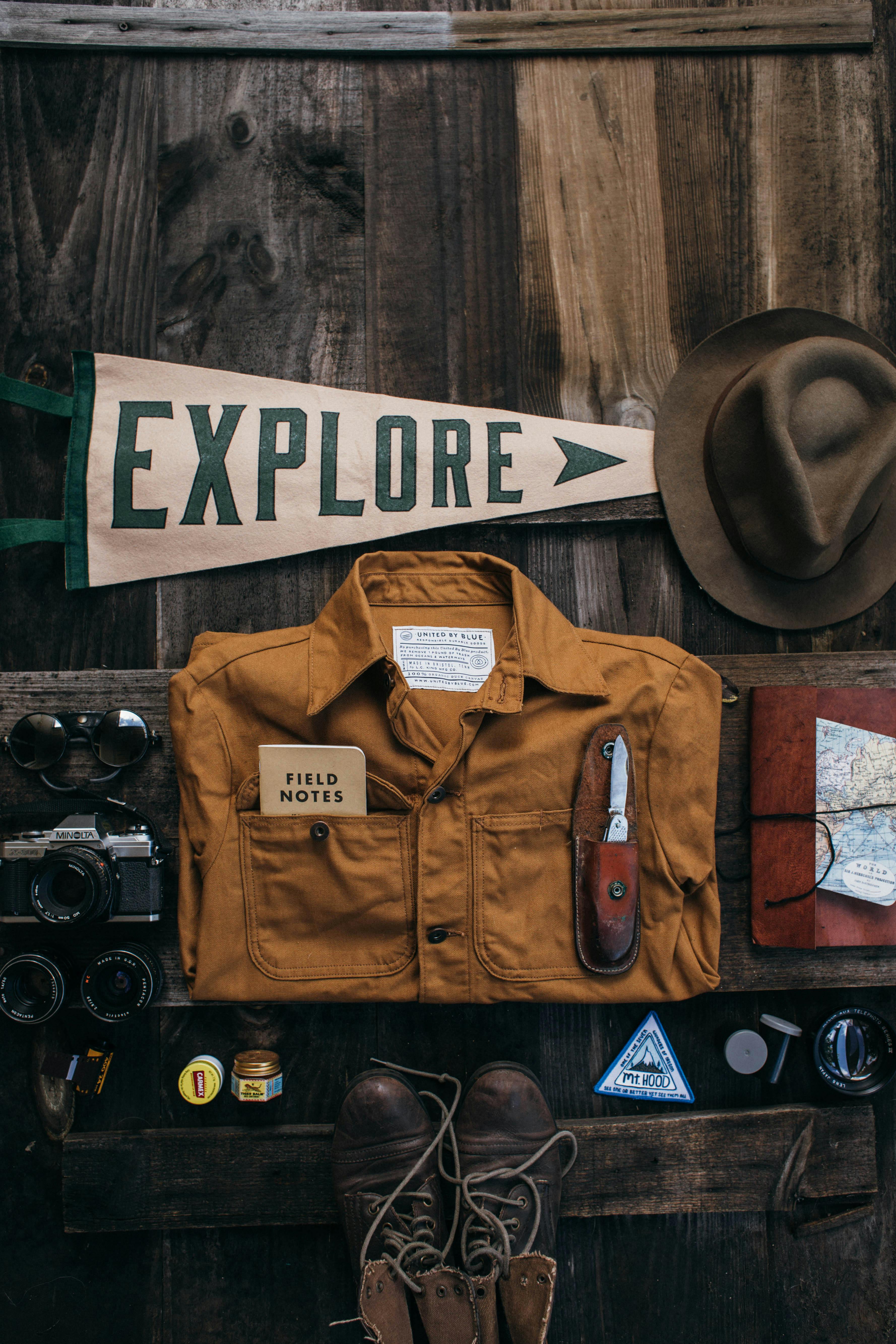 Overhead of vintage photo camera near lenses and sunglasses near shoes and jacket near notebook with map and knife near hat and poster with word explore placed on table