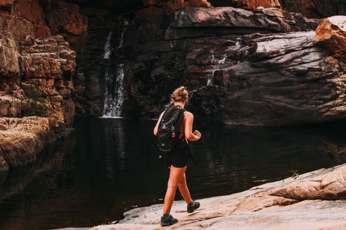 Free Back view full length of unrecognizable lady in backpack and casual outfit walking near small waterfall in rocky mountain near lake in canyon in daytime Stock Photo