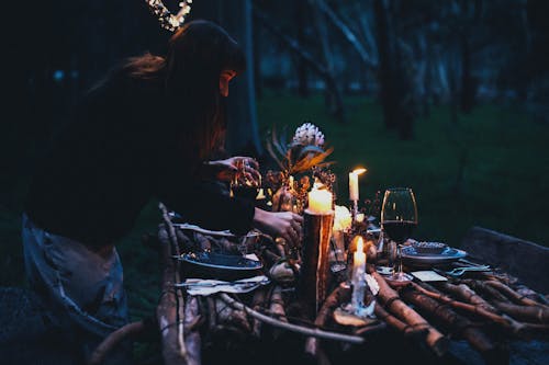 Side view of unrecognizable lady serving wineglasses on table with plates and forks with knifes near candles in evening in nature near forest