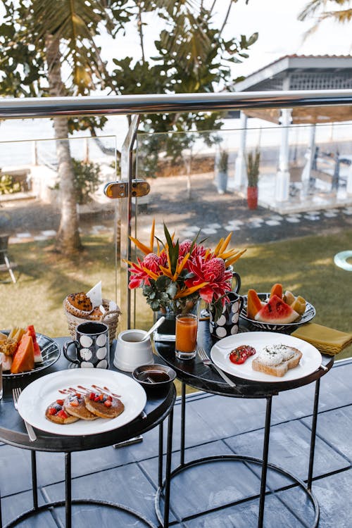 Free Round tables on balcony with pancakes with berries near plate with sandwich with poached eggs and tomatoes and plates with different fruits near mugs with drink and vase with flowers Stock Photo