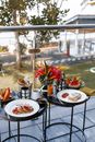 Round tables on balcony with pancakes with berries near plate with sandwich with poached eggs and tomatoes and plates with different fruits near mugs with drink and vase with flowers