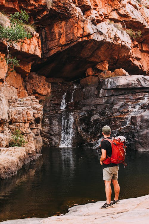Back view full body of faceless man in casual clothes and backpack admiring small waterfall in rocky hill near river in summer day in canyon