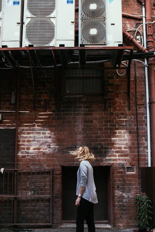 Side view of unrecognizable female with flying hair standing on street near aged house with air conditioning units in city