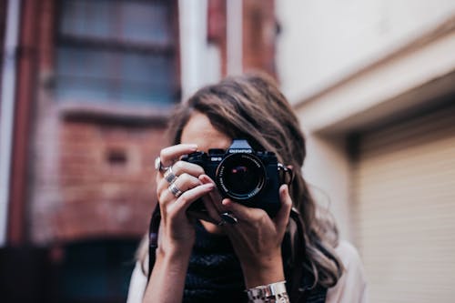 Unrecognizable female photographer taking photo and covering face with camera while standing on street against brick building on blurred background