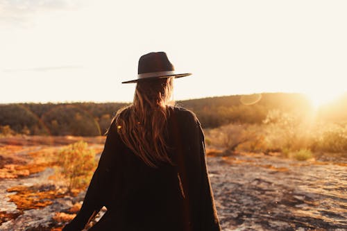 Back view of anonymous female in black headwear standing on rocky ground against coniferous forest during sunset time in nature
