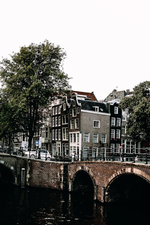 Street of city with houses and trees placed near old stone bridge with arched elements near calm river against cloudless sky