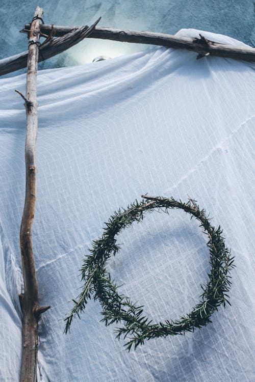 Rural design of green round coniferous wreath hanging on wall on white fabric with construction of wooden sticks in room