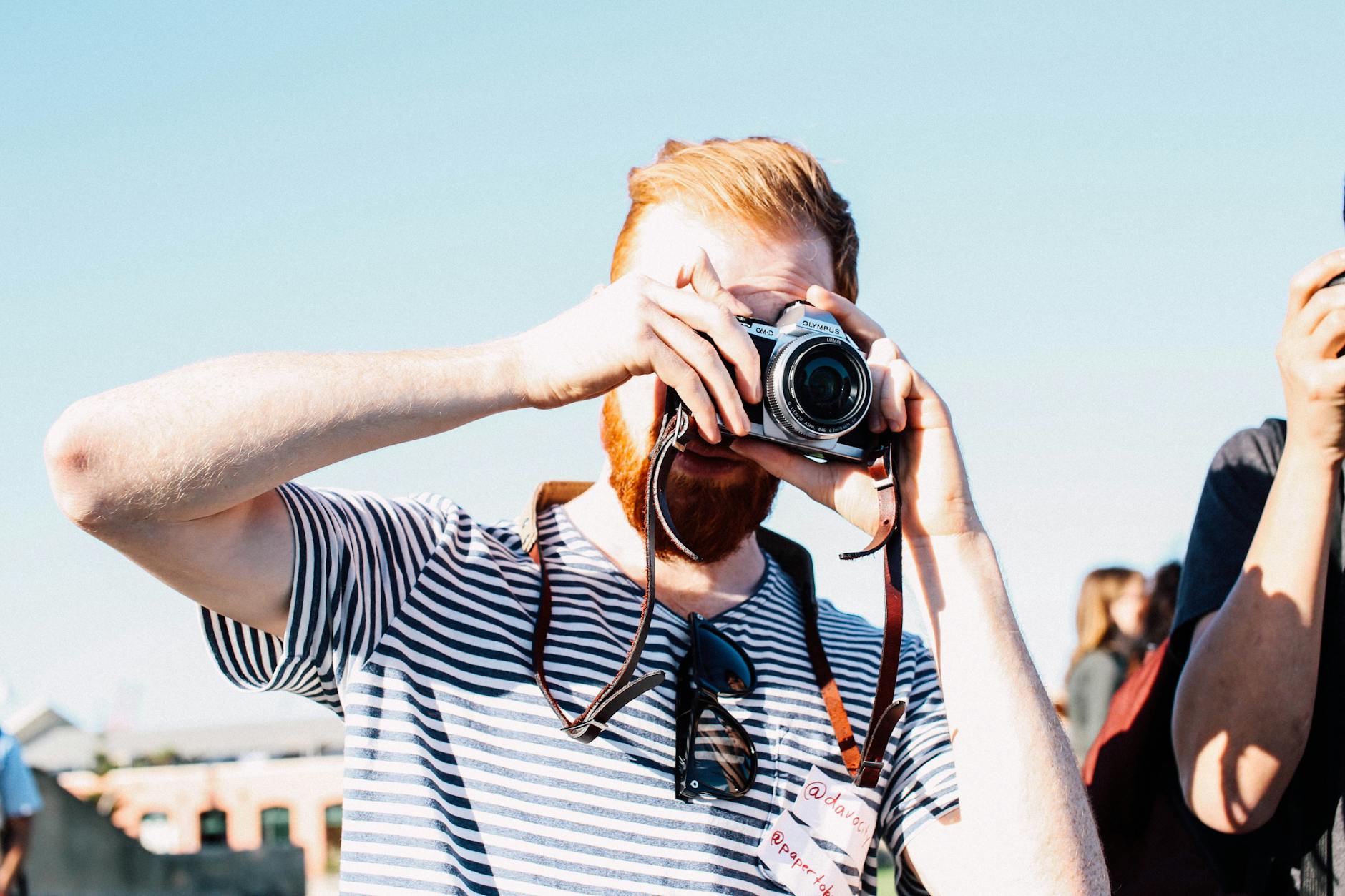 Unrecognizable redhead bearded male photographer in casual outfit concentrating on taking picture by vintage photo camera on street event at