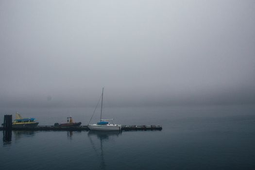 Lonely boats moored near small pier surrounded calm sea with misty horizon line on overcast day