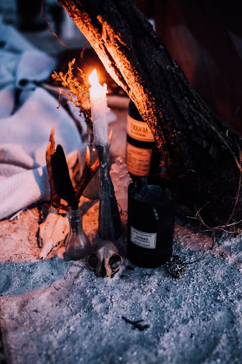 Free From above of gloomy composition with bottle of alcohol and skull near burning candle and bottles placed on white sand near peace of wood Stock Photo