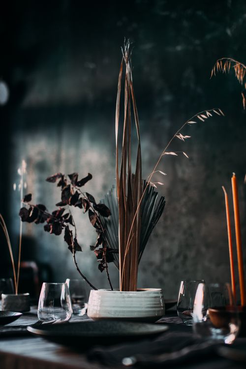 Free Composition of dry leaves and branches in white vase placed on table with dark tablecloth and utensil among candles and glasses with dark wall on background Stock Photo